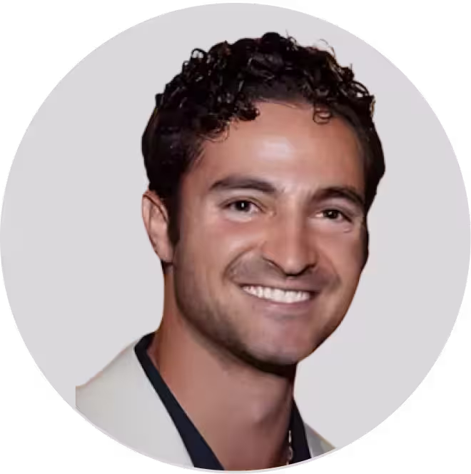 Smiling man with curly dark hair wearing a light-colored jacket over a dark shirt against a plain background.